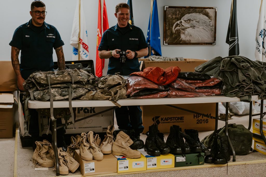 Two men in U.S. Coast Guard uniforms stand behind a table piled with gear.