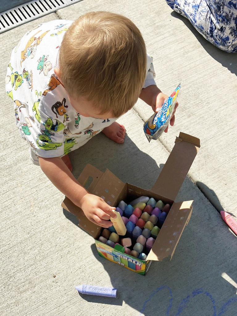 A toddler crouches on concrete, choosing their color of chalk to draw with.