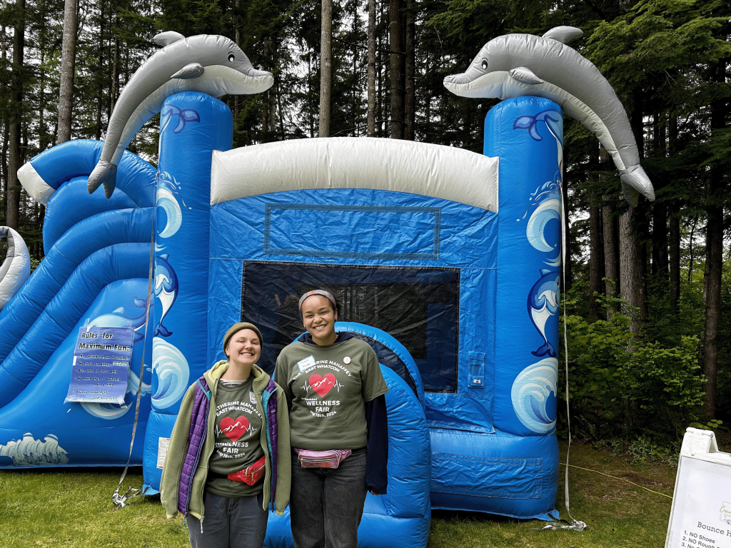 Two people smile in front of a big blue aquatic-themed bouncy house that has wave shapes and inflatable dolphins.