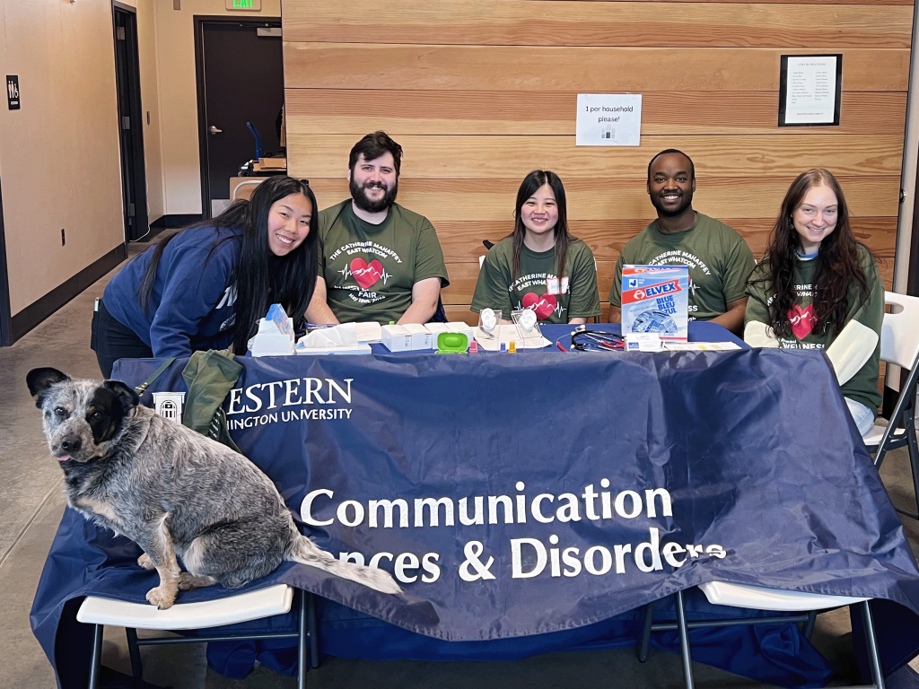 Five people wearing army green "The Catherine Mahaffey East Whatcom Wellness Fair" with beating heart clipart on them. Their info table says "Western Washington University Communication Sciences & Disorders." A dog sits on a chair in front of the table.