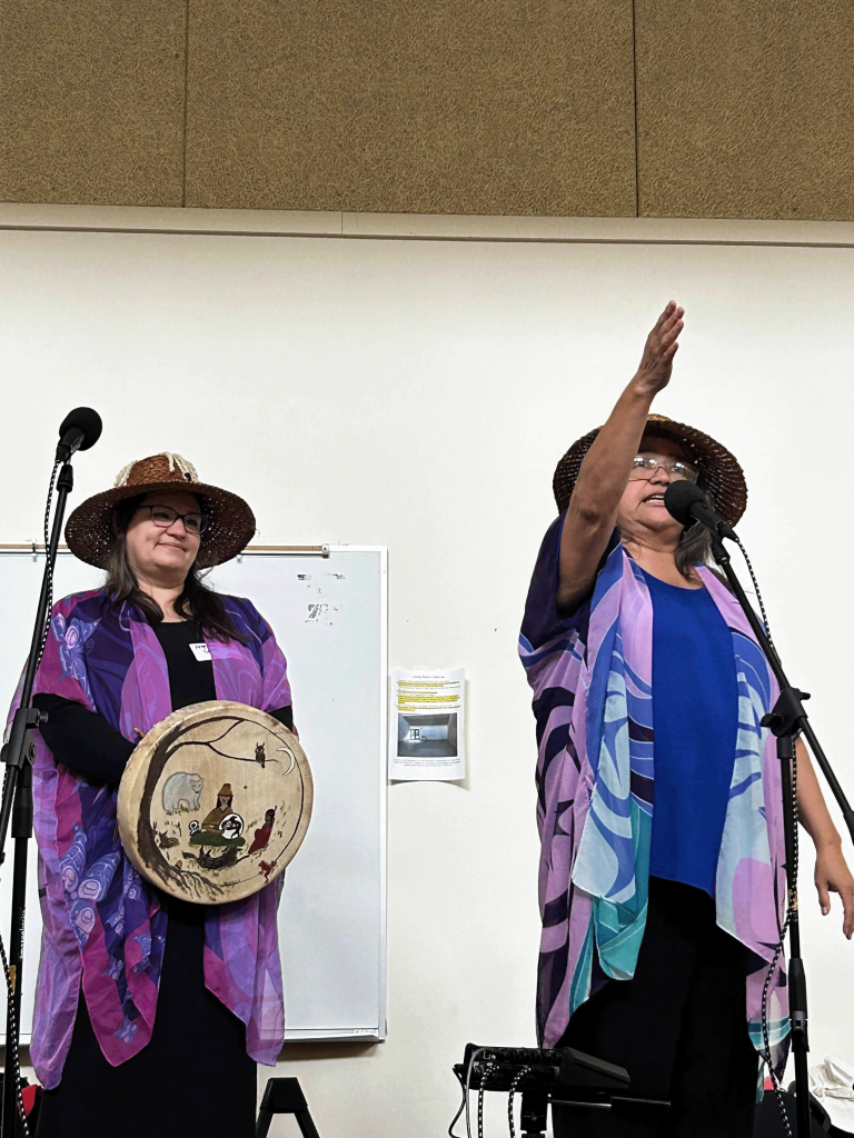 Two people in Native American clothing, one holding a Native American drum, stand on a stage behind microphones.