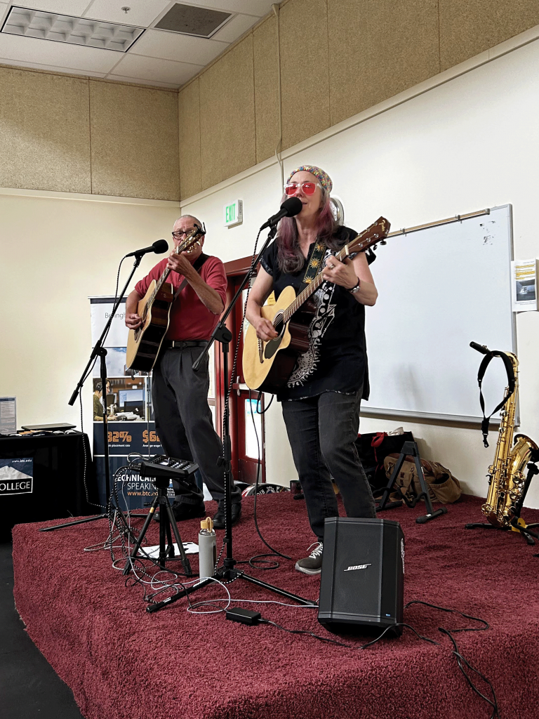 Two people stand on a stage, holding guitars and singing into microphones.