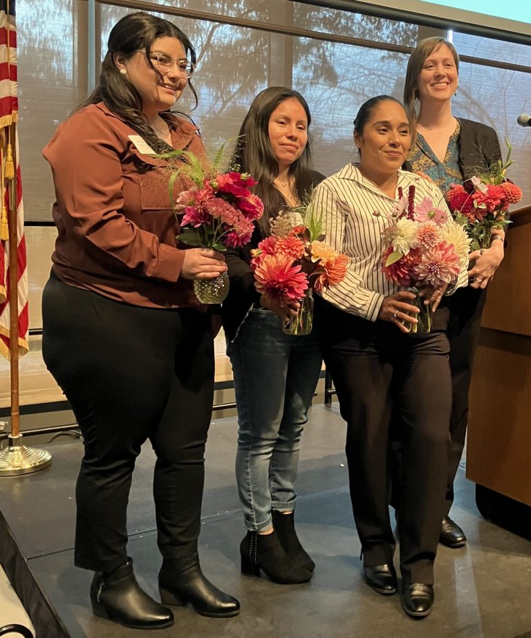 Five people stand on a stage next to a podium, smiling and holding vibrant bouquets of flowers.