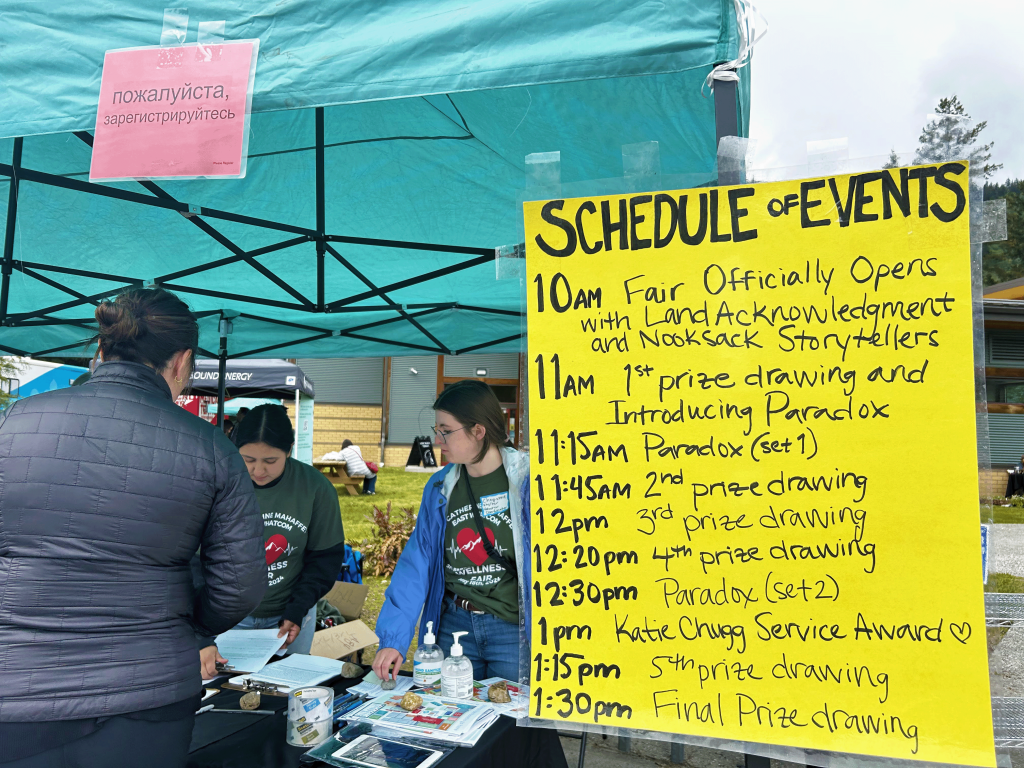 On a bright yellow poster, a "schedule of events" is written. Three people stand under a teal canopy next to it at their info table.