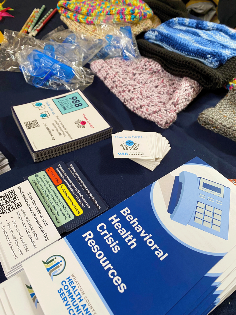 An info table contains knitted hats and resources, including a pamphlet from the Whatcom County health department called "Behavioral Health Crisis Resources" and handouts about the 988 suicide & crisis lifeline.