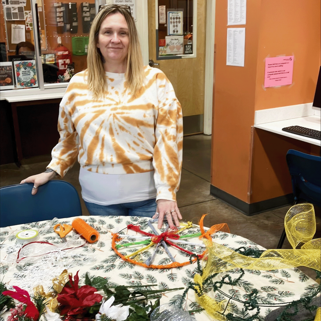 A woman wearing a tie-dye sweatshirt stands in front of a circular table with crafting materials on it and a holiday wreath she's been working on.