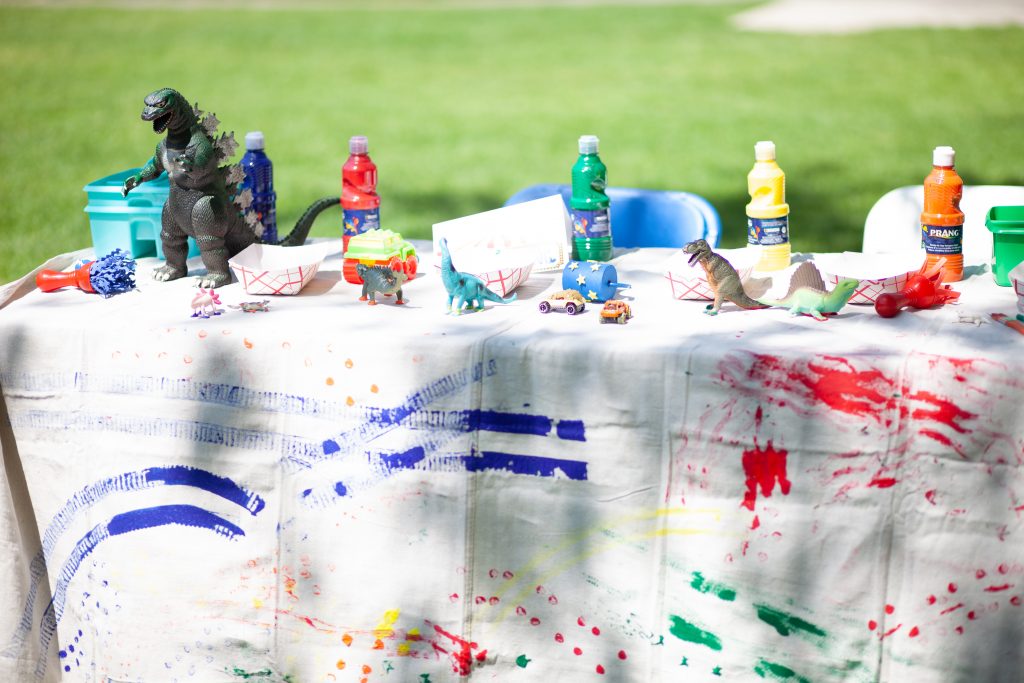 A table covered in a tablecloth smeared with paint; on it, paints, toy cars, and dinosaurs.