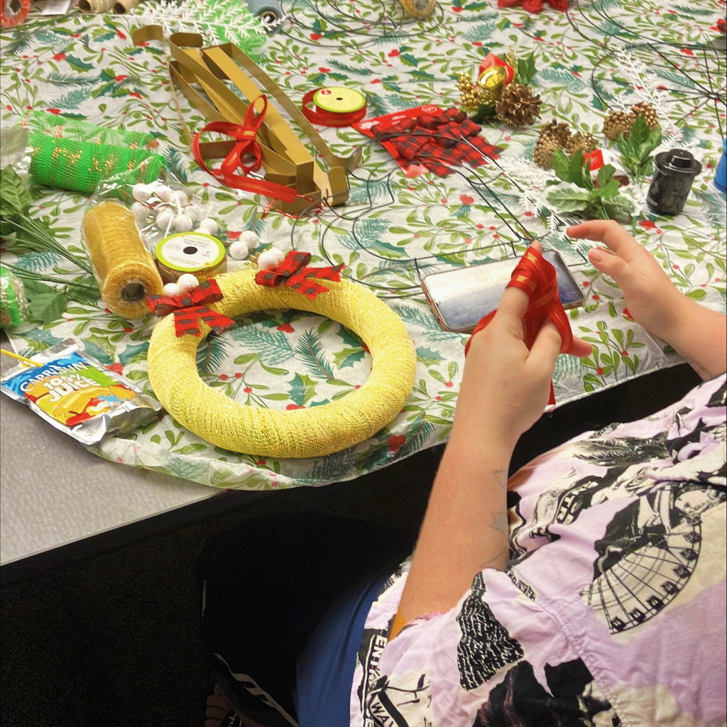 A person sits at a table with a tablecloth and crafting supplies on it, making their own holiday wreath.