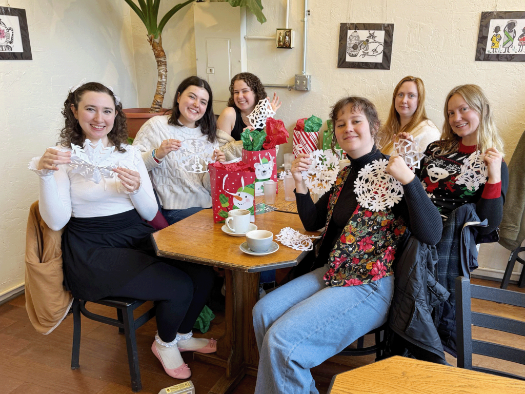 A group of people sit together at a table with gift bags and warm beverages on it, showing off the paper snowflakes they made together.